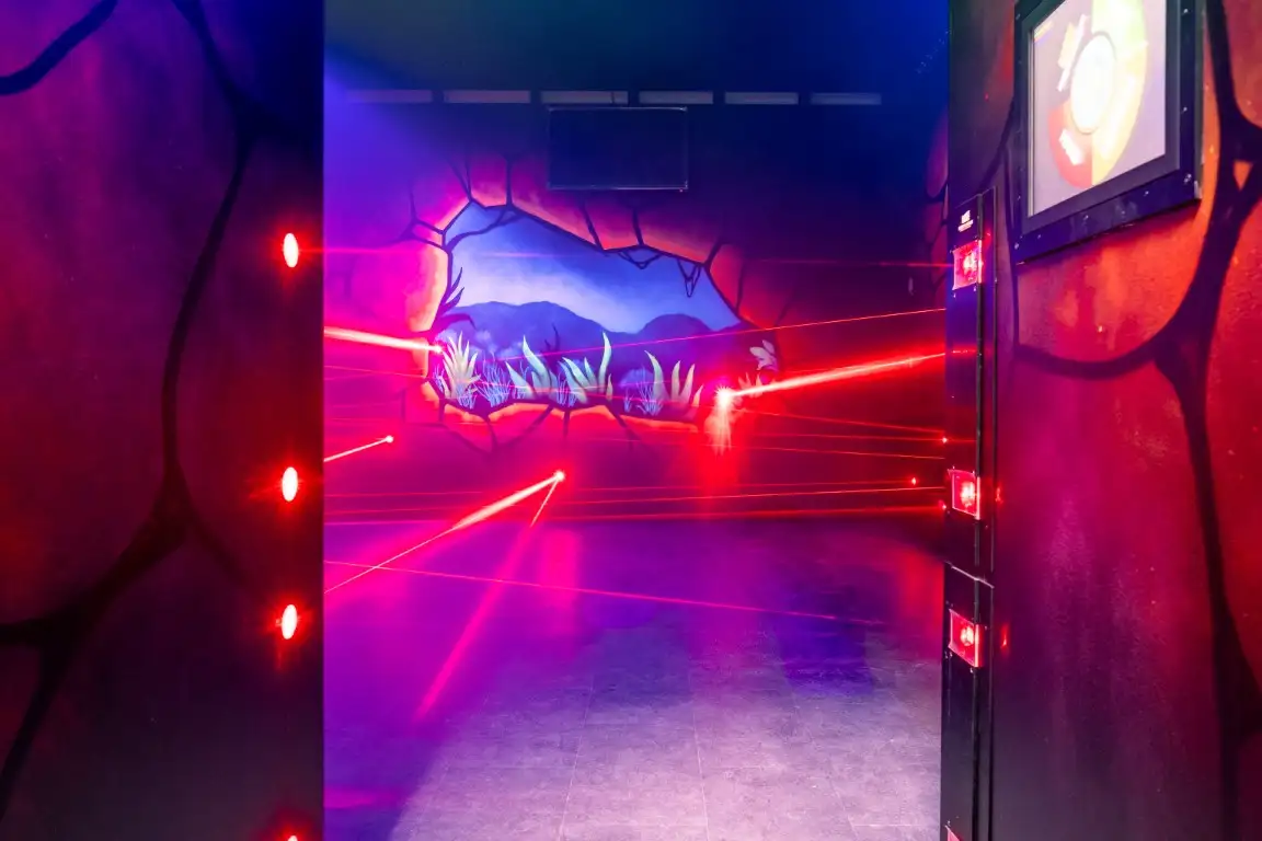Blick durch den Eingang in den Laser Maze: rote Strahlen kreuzen den Raum, an den Wänden leuchtende Felsmalereien.